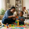 A warm, cheerful Nairobi nanny sitting on a colourful play mat with a toddler, both laughing and playing with building blocks — the child looks relaxed and engaged, with a bright, well-furnished living room in the background, conveying trust, safety, and a growing bond between child and caregiver